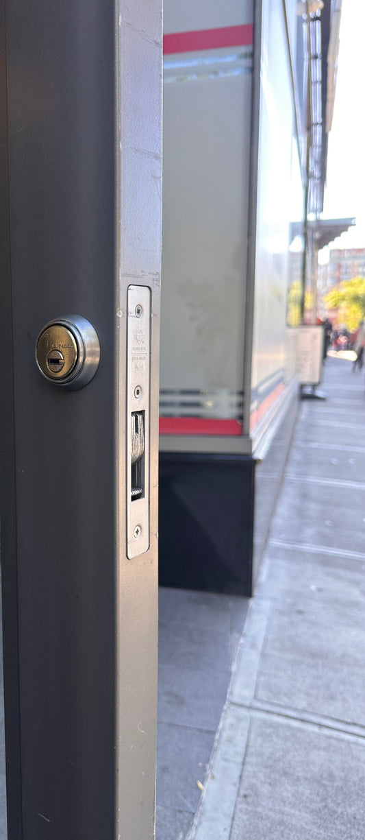 Adams Rite faceplate and mortise cylinder on an aluminum storefront door — the lock a keyless SK1850S(sfic) swap replaces in the same pocket
